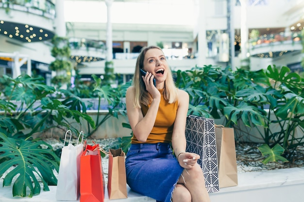 Jonge vrouw consument in het winkelcentrum bladert door chat en gebruikt het gebruik van een smartphone. vrouw staat met een mobiele telefoon in haar handen in het winkelcentrum. binnen. gelukkig shopper meisje met cadeauzakjes aankopen doen