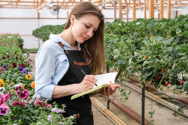 Jonge vrouw die aantekeningen maakt in een notitieboekje terwijl ze in een industriële kas werkt