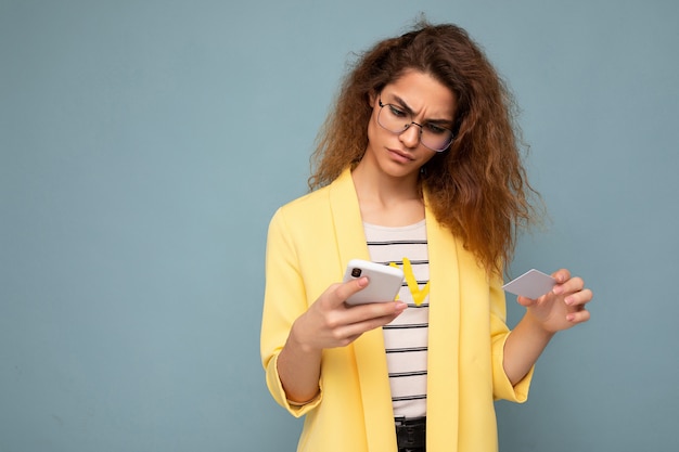 Jonge vrouw die alledaagse kleding draagt, geïsoleerd over achtergrond met telefoon en creditcard die online winkelen via creditcard betaalt en naar het smartphonescherm kijkt.