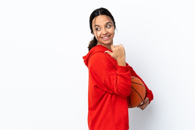 Jonge vrouw die basketbal over geïsoleerde witte muur speelt die naar de kant wijst om een product te presenteren