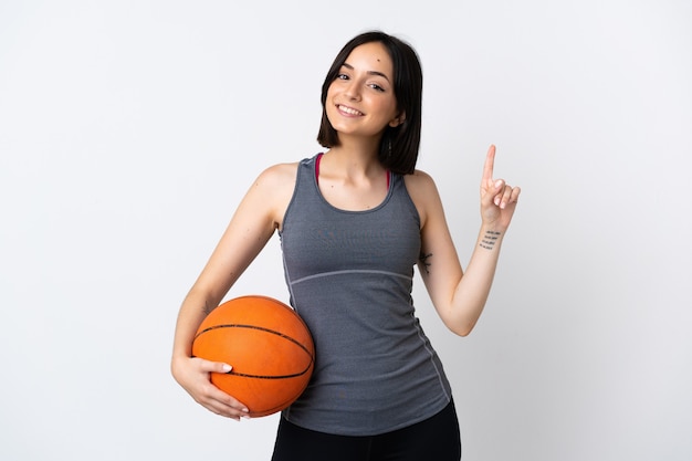 Jonge vrouw die basketbal speelt dat op witte muur wordt geïsoleerd die en een vinger in teken van het beste toont opheft
