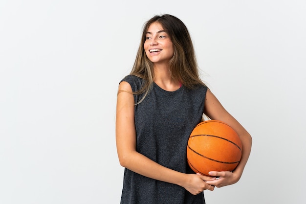 Jonge vrouw die basketbal speelt geïsoleerd op een witte achtergrond, naar de zijkant kijkt en glimlacht