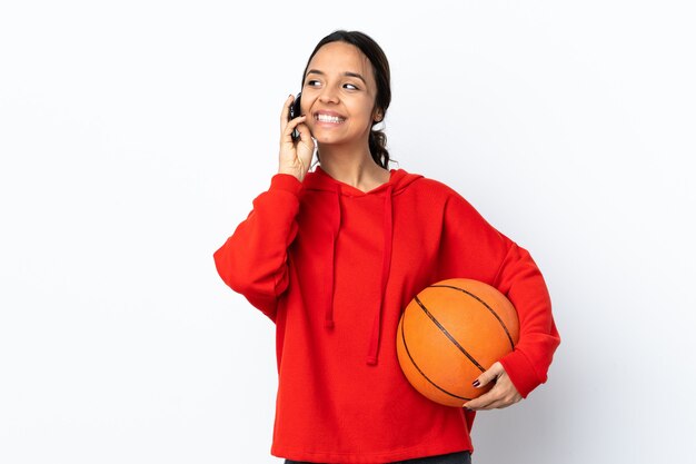 Jonge vrouw die basketbal speelt over een geïsoleerde witte achtergrond die een gesprek voert met de mobiele telefoon