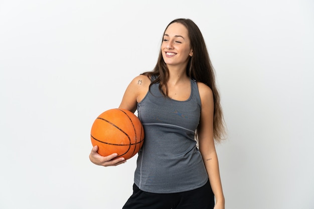 Jonge vrouw die basketbal speelt over een geïsoleerde witte achtergrond die naar de zijkant kijkt en glimlacht