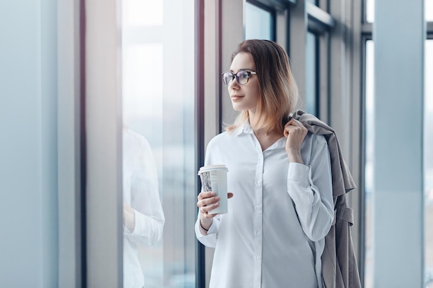 Jonge vrouw die bij het raam van een modern zakencentrum staat met een kopje koffie