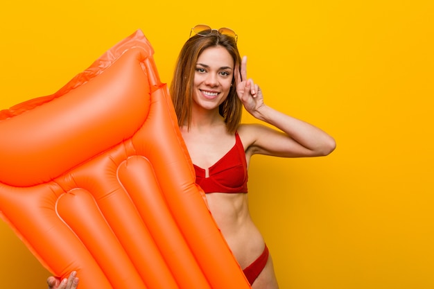 Jonge vrouw die bikini draagt, een luchtmatrasbed houdt dat overwinningsteken toont en breed glimlacht.