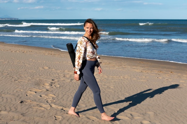Jonge vrouw die blootsvoets op het strand loopt met een mat over haar schouder