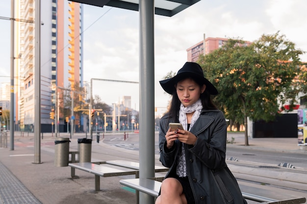 Jonge vrouw die celtelefoon kijkt die de bus wacht