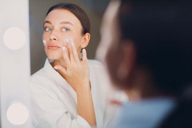 Jonge vrouw die cosmetische witte crème op haar gezicht aanbrengt in de spiegel