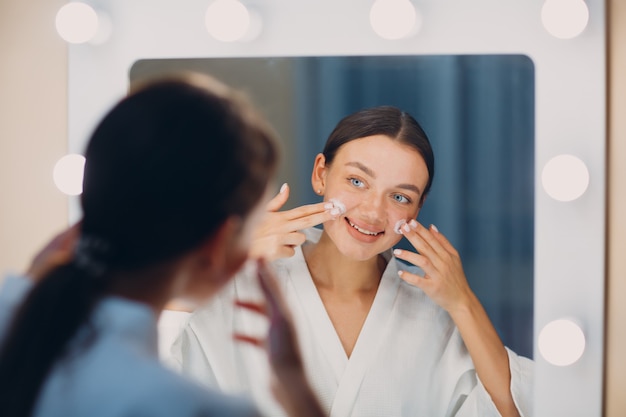 Jonge vrouw die cosmetische witte crème op haar gezicht aanbrengt in de spiegel