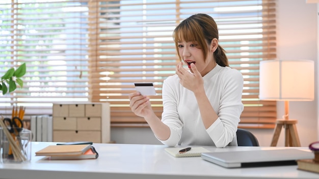 Jonge vrouw die creditcard vasthoudt en bekijkt met opgewonden en verrast Online winkelen internetbankieren concept