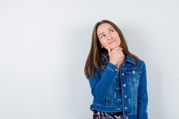 Jonge vrouw die de vinger op de kin houdt, omhoog kijkt in een spijkerjasje, jurk en peinzend kijkt. vooraanzicht.