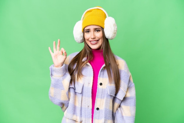 Jonge vrouw die de wintermof draagt over geïsoleerde chroma key achtergrond die ok teken met vingers toont