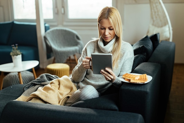 Jonge vrouw die digitale tablet gebruikt terwijl ze koffie drinkt en thuis op de bank ontspant