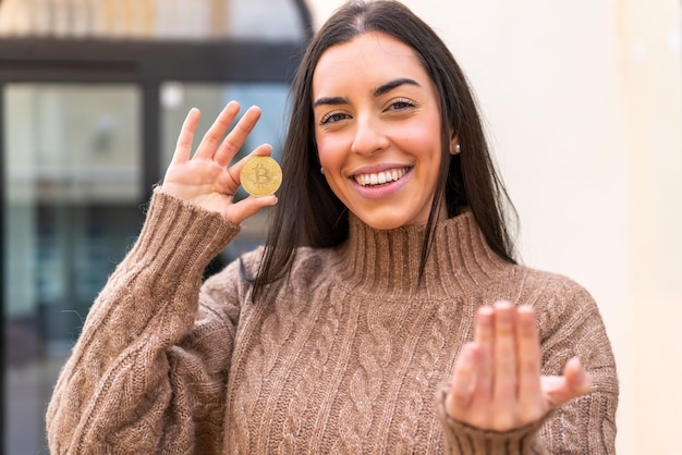 Jonge vrouw die een bitcoin in de open lucht vasthoudt en uitnodigt om met de hand te komen blij dat je gekomen bent