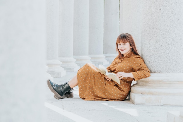 Jonge vrouw die een boek leest en naar de camera glimlacht terwijl ze op de vloer zit op een architectonische plek. Gelukkig tijdens het leren en lezen van avonturen. Ruimte kopiëren. jaren 80 sfeer. Trendy stijl jurk