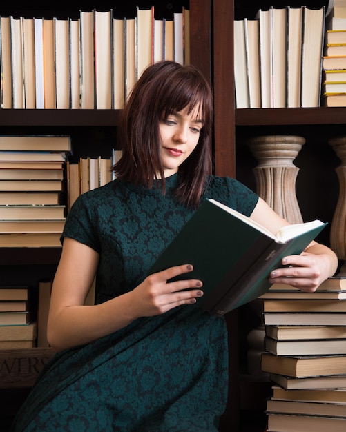 Jonge vrouw die een boek leest terwijl ze tegen een boekenplank in de bibliotheek zit