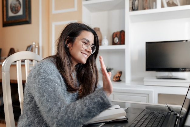 Jonge vrouw die een bril draagt die met haar hand zwaait tijdens een videogesprek