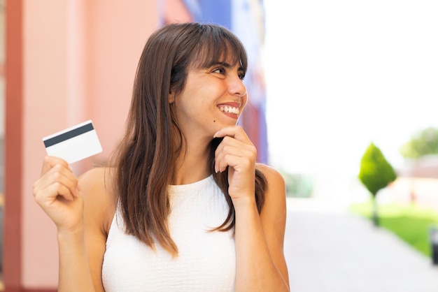 Jonge vrouw die een creditcard vasthoudt en buitenshuis een idee denkt en naar de kant kijkt