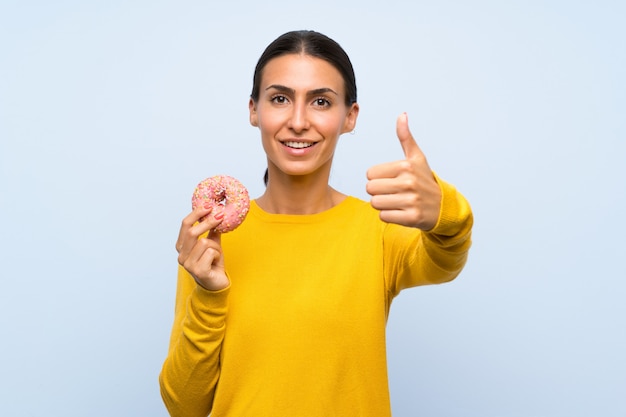 Jonge vrouw die een doughnut over geïsoleerde blauwe muur met omhoog duimen houdt omdat iets goeds is gebeurd