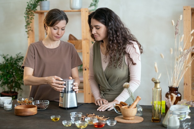 Jonge vrouw die een elektrisch keukenapparaat gebruikt voor het malen van ingrediënten voor handgemaakte zeep en haar vriend laat zien op welke knop ze moet drukken