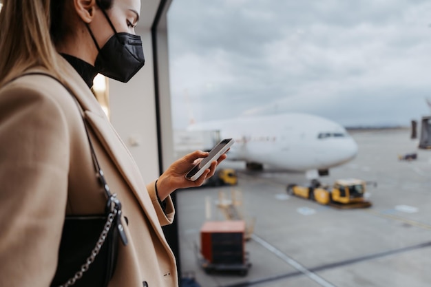 Jonge vrouw die een gezichtsmasker draagt en een mobiele telefoon vasthoudt Mobiele app gebruiken op de luchthaven