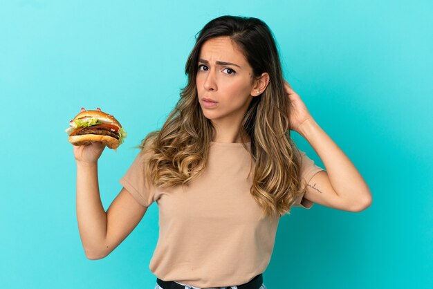 Jonge vrouw die een hamburger over geïsoleerde achtergrond houdt die twijfels heeft