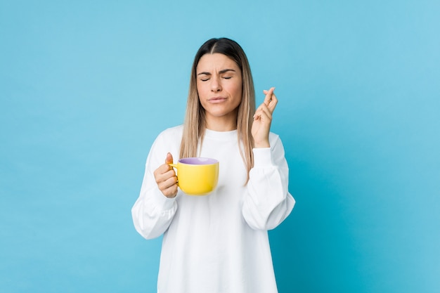 Jonge vrouw die een koffiekop houdt die vingers kruist voor het hebben van geluk