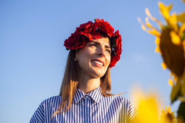 Jonge vrouw die een krans van rode bloemen draagt die van de zonsondergang op een zonnebloemgebied genieten.