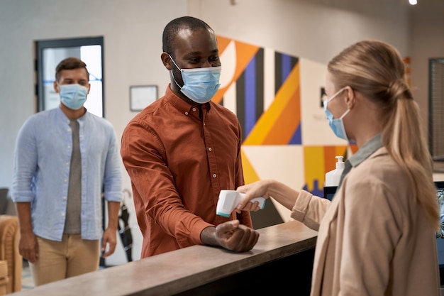 Jonge vrouw die een medisch gezichtsmasker draagt dat bij de receptie op kantoor staat en infrarood gebruikt