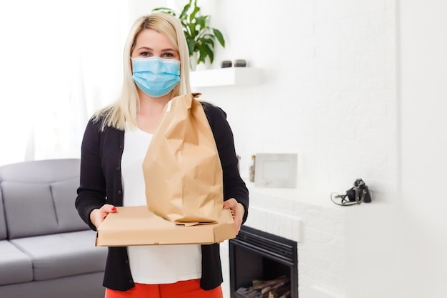 Jonge vrouw die een medisch masker draagt en een pakket ontvangt van de bezorger binnenshuis. Preventie van virusverspreiding