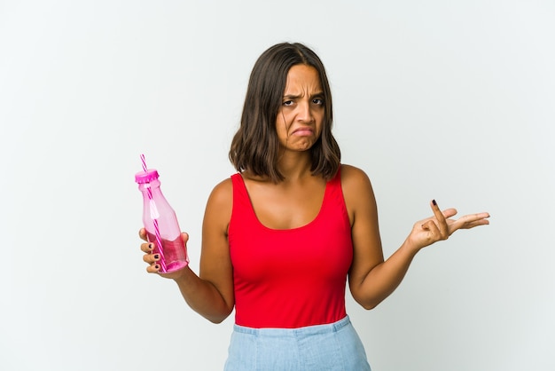 Jonge vrouw die een milkshake houdt die op witte muur wordt geïsoleerd die twijfelt en schouders ophaalt in vragend gebaar