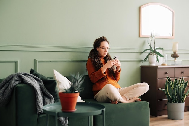 Jonge vrouw die een mobiele telefoon gebruikt terwijl ze thuis op de bank zit