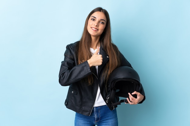 Jonge vrouw die een motorhelm houdt die op blauwe muur wordt geïsoleerd die duimen op gebaar geeft