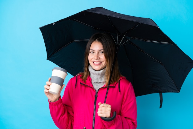 Jonge vrouw die een paraplu en een koffie houdt om over geïsoleerde blauwe muur weg te halen