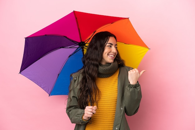 Jonge vrouw die een paraplu houdt die op roze achtergrond wordt geïsoleerd die naar de kant wijst om een product te presenteren