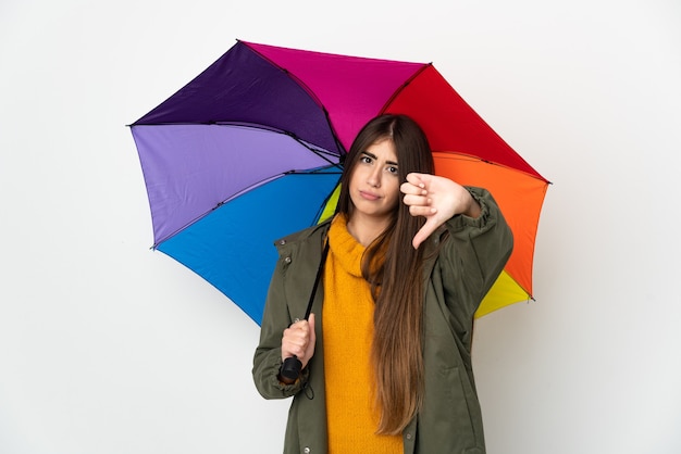 Jonge vrouw die een paraplu houdt die op witte muur wordt geïsoleerd die duim met negatieve uitdrukking toont