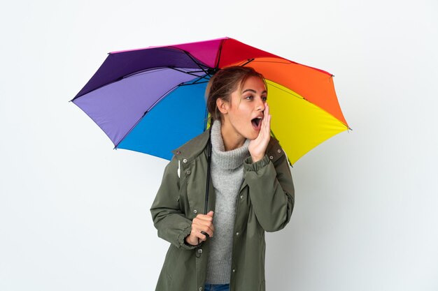 Jonge vrouw die een paraplu houdt die op witte muur wordt geïsoleerd die met wijd open mond aan de kant schreeuwt