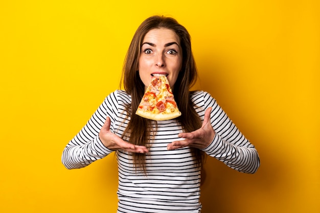 Jonge vrouw die een plak van hete verse pizza met haar tanden op geel houdt.