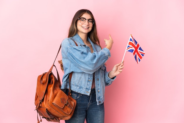 Jonge vrouw die een vlag van het Verenigd Koninkrijk houdt die op roze muur wordt geïsoleerd die terug richt
