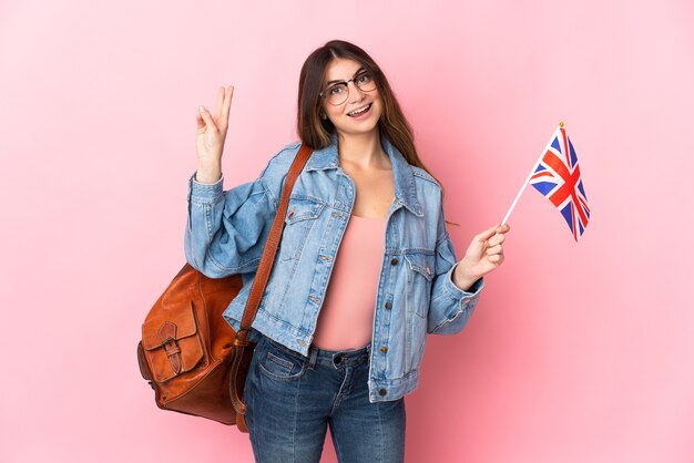 Jonge vrouw die een vlag van het Verenigd Koninkrijk houdt die op roze wordt geïsoleerd die en overwinningsteken glimlacht toont