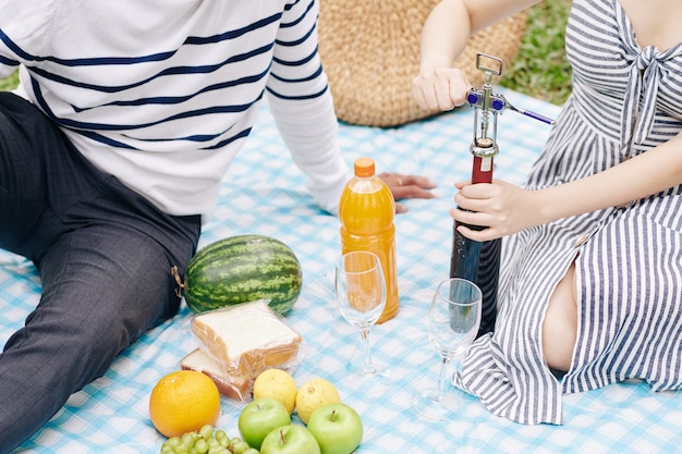 Jonge vrouw die een vleugelkurkentrekker gebruikt bij het openen van een fles rode wijn bij een picknick in het park