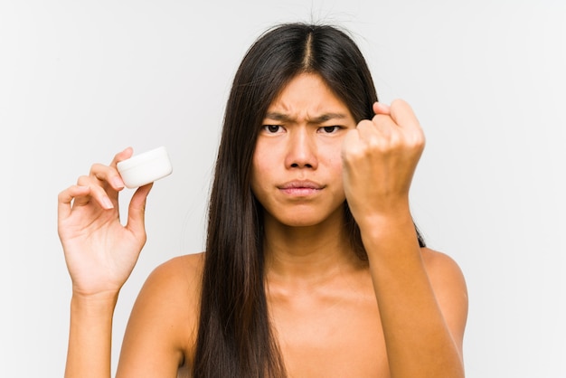 Jonge vrouw die een vochtinbrengende crème houdt die vuist toont aan camera, agressieve gelaatsuitdrukking.