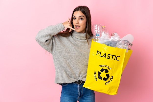 Jonge vrouw die een zak vol plastic flessen houdt om te recyclen geïsoleerd op roze muur die telefoongebaar maakt. Bel me terug teken