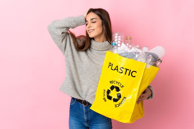 Jonge vrouw die een zak vol plastic flessen houdt om te recyclen geïsoleerd op roze muur die veel glimlacht