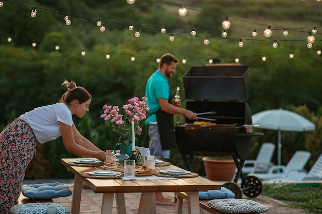 Foto jonge vrouw die eettafel dekt terwijl haar man naast haar aan het barbecueën is