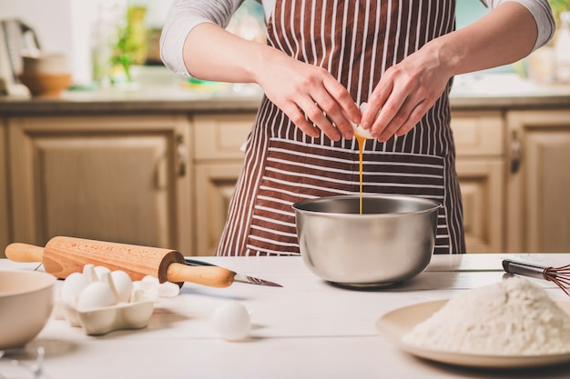 Jonge vrouw die ei over kom met deeg breekt, close-up. Een vrouw in een gestreept schort kookt in de keuken