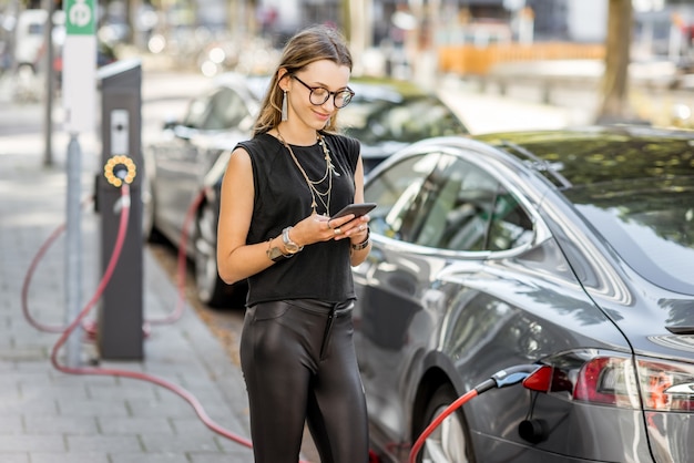 Jonge vrouw die elektrische auto oplaadt die met smartphone buiten op straat staat in de stad Rotterdam