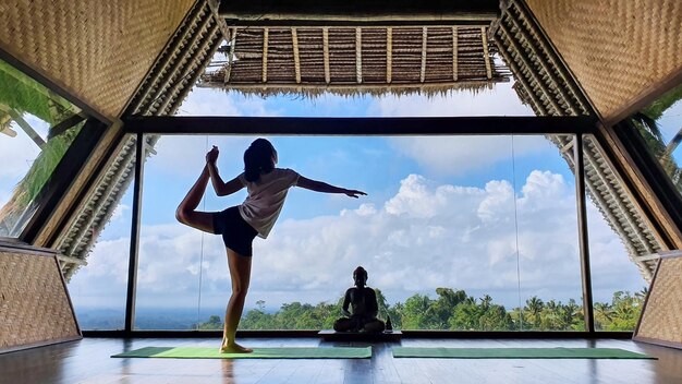 Jonge vrouw die evenwicht doet terwijl ze yoga beoefent in de buurt van een Boeddhabeeld met blauwe hemelachtergrond in de ochtend