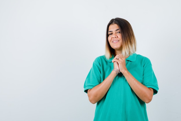 Jonge vrouw die geklemde handen toont in een smekend gebaar in polot-shirt en er dankbaar uitziet, vooraanzicht.